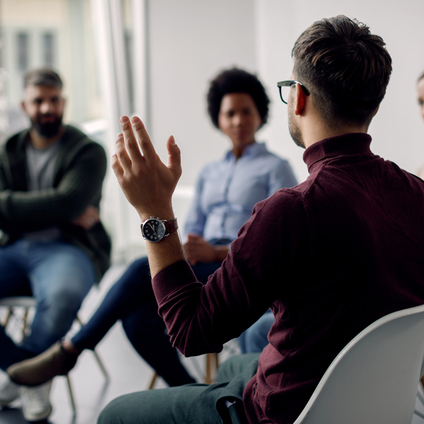 Rear view of man raising his hand to ask a question while attending group therapy.